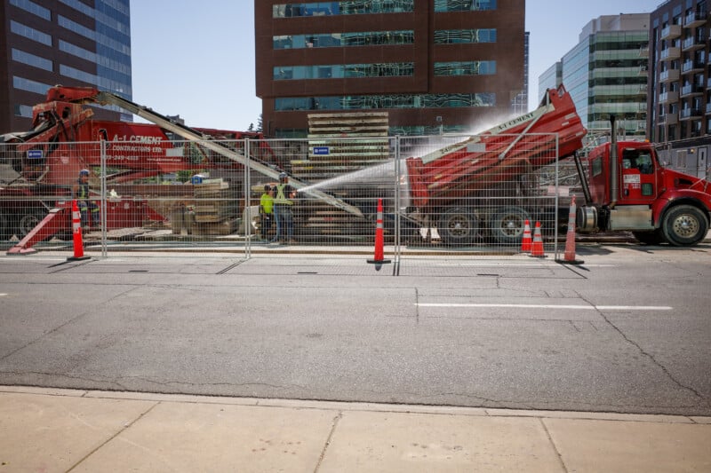 Construction site on a city street with workers, orange cones, a red cement truck, and a water hose spraying over materials. Office buildings are visible in the background behind a metal fence.