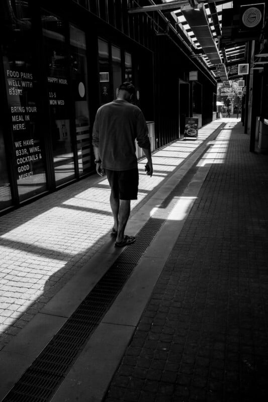 A man in casual clothes and flip-flops walks alone down a sunlit, shadowy corridor between buildings. The walkway is lined with windows and signs, and the scene has a tranquil, contemplative mood.