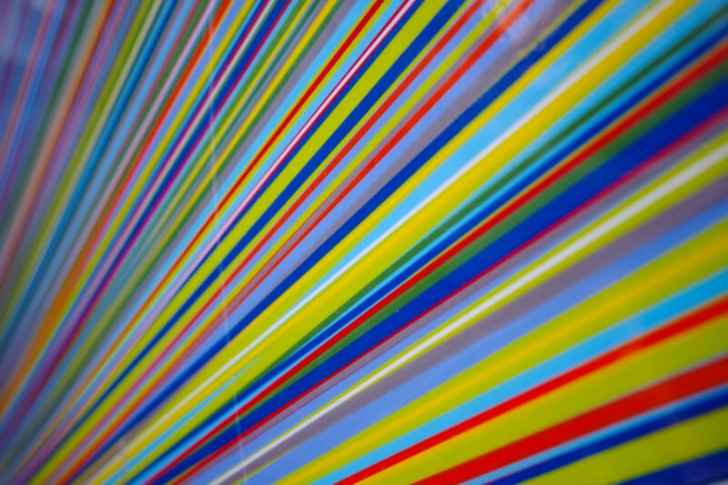 Colorful, diagonal lines of varying thickness in blue, red, yellow, green, and purple create a vibrant, abstract pattern across the image, converging toward one corner on a light background.