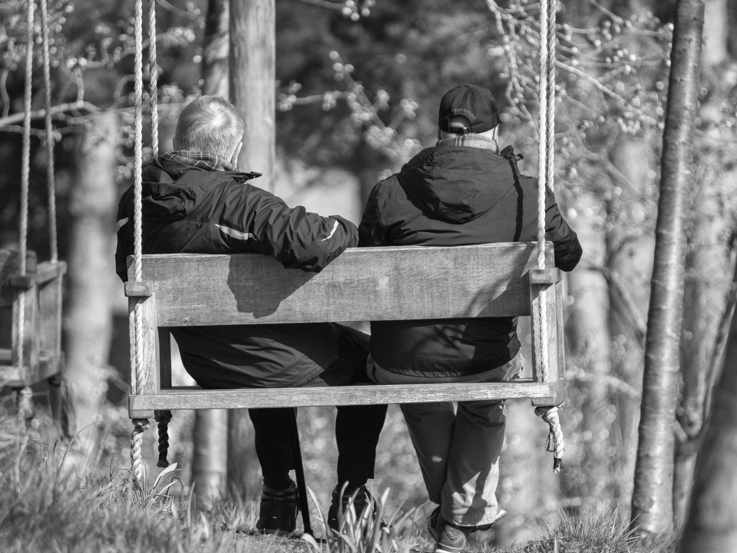 Two people in jackets sit side by side on a wooden swing outdoors, facing away from the camera. The background features trees and sunlight, creating a peaceful and relaxed atmosphere.
