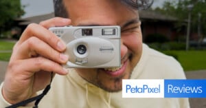 A smiling person in a light hoodie holds a silver RETO Ultra Wide & Slim film camera to their face. The PetaPixel Reviews logo is overlaid in the bottom right corner. A building and trees are blurred in the background.