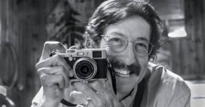 A smiling man with glasses and a mustache holds a vintage camera up to his face indoors. The black-and-white photo captures a joyful, candid moment.