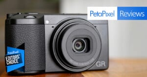 A compact black digital camera sits on a wooden surface. The lens reads "GR LENS f=18.3mm 1:2.8." Overlay text reads "PetaPixel Reviews" and "EDITORS' CHOICE" in a blue badge.