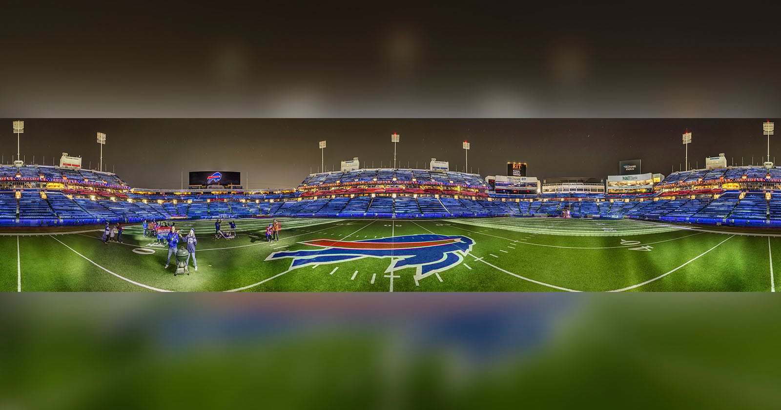 A wide-angle view of an empty football stadium at night, with bright lights and a large buffalo logo at midfield. A few people stand on the turf near the center. Blue seats fill the stands in the background.