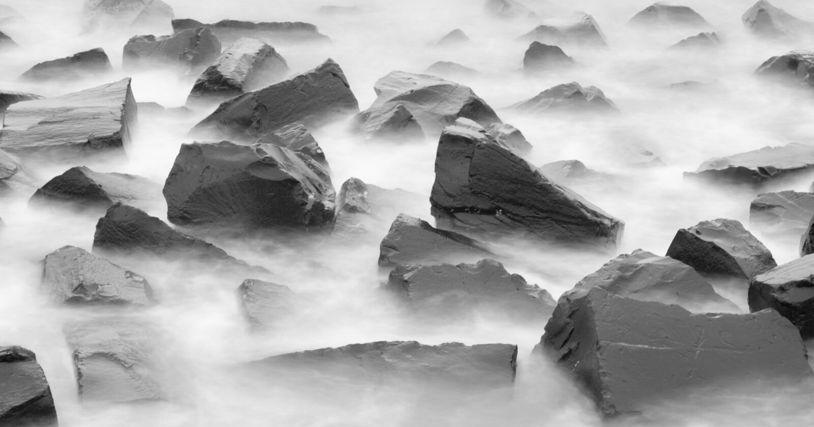 Large, jagged rocks partially submerged in misty, flowing water, creating a surreal and ethereal scene in black and white.