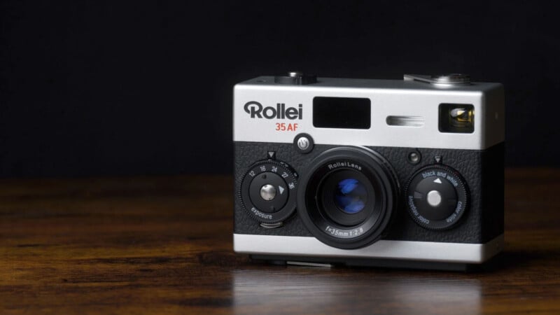 A vintage Rollei 35 AF film camera with a silver and black body is placed on a wooden surface against a dark background.