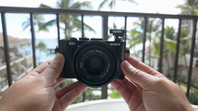 A person holds a Sony RX1 camera with a Carl Zeiss lens in front of a balcony railing, with palm trees and a blurred outdoor tropical background visible.