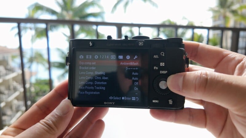 A person holds a Sony digital camera, adjusting settings on the screen. In the background, blurred palm trees and a balcony railing suggest a tropical outdoor location.