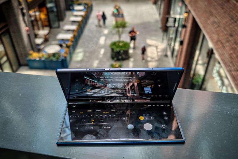A foldable smartphone in camera mode rests on a railing, capturing an image of a modern, outdoor shopping area below with a few people walking and plants along the walkway.