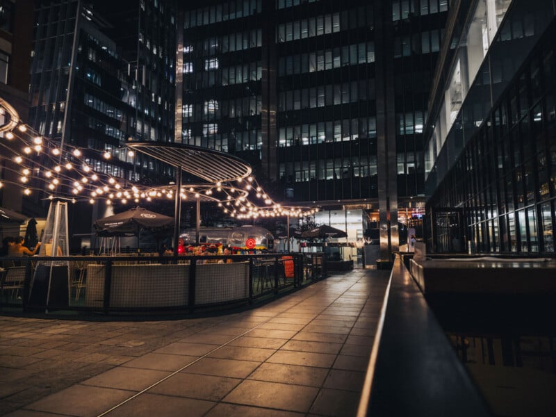 Outdoor patio with string lights, empty tables, and chairs at night, surrounded by tall modern office buildings with illuminated windows. The scene appears calm and urban, with soft warm lighting from the decorations.