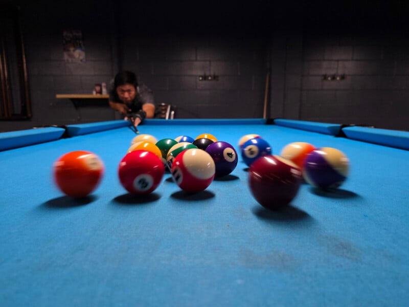 A person is lining up a pool shot, with billiard balls scattering across a bright blue pool table in the foreground. The image is slightly blurred, suggesting motion.