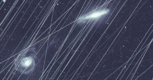 A night sky filled with stars and galaxies is crossed by numerous bright, straight streaks of light, likely caused by satellite trails, partially obscuring the celestial objects.