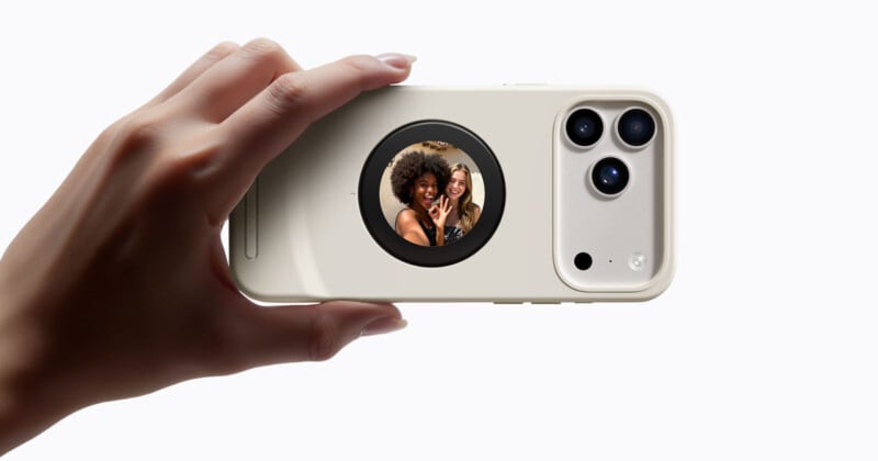 A hand holds a smartphone with a beige case displaying a small circular screen on the back, showing two women smiling and making peace signs for a selfie.