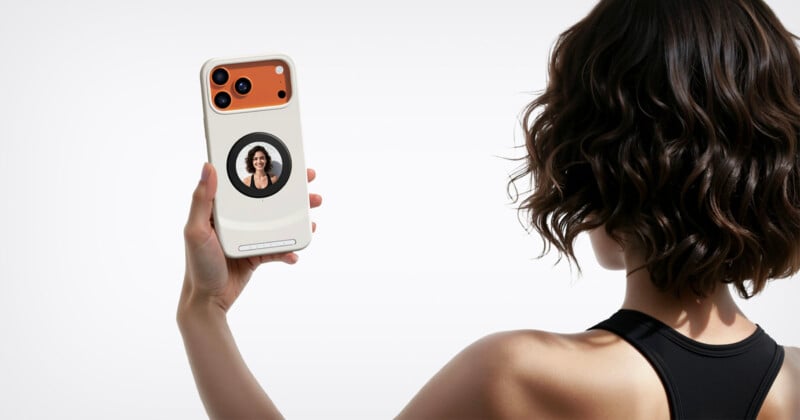 A woman with short, wavy hair and a black sleeveless top holds up a smartphone with a circular display on the back showing her own image, as if taking a selfie.