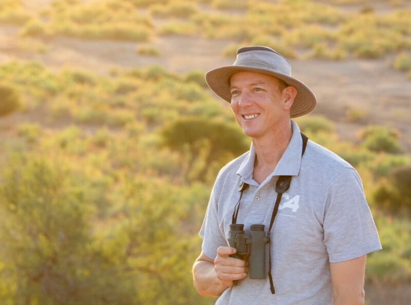 A smiling person in a gray hat and polo shirt stands outdoors, holding binoculars with a sunlit grassy landscape in the background.