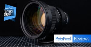 A camera lens is displayed on a checkered surface with a "Petapixel Editors' Choice" badge and "Petapixel Reviews" text in the bottom right corner. The background is dark and out of focus.