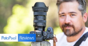 A man with a beard stands behind a camera with a large lens mounted on a wooden post. The image features a "PetaPixel Reviews" graphic overlay in the lower left corner. The background is blurred greenery.