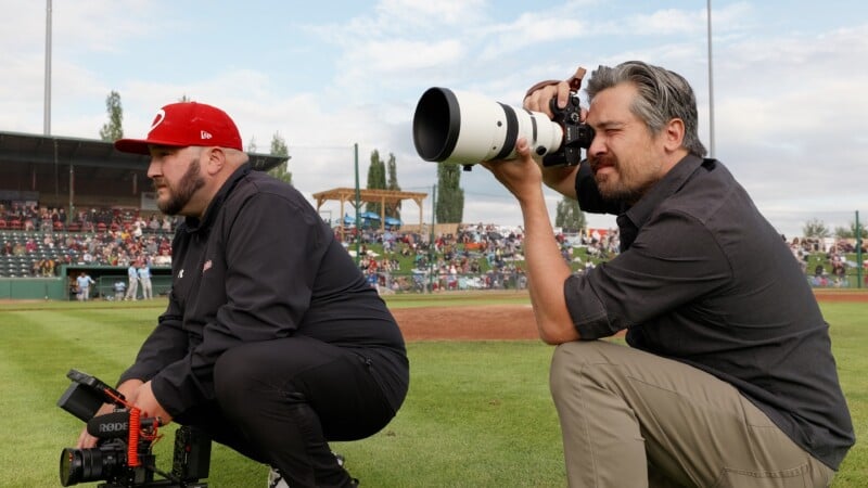 Two men kneel on a baseball field; one holds a video camera, the other takes photos with a large telephoto lens. A stadium crowd and players are visible in the background.