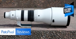A large white telephoto camera lens is resting on a concrete surface. The image features "PetaPixel Reviews" and "Editor's Choice" badges in the corners.