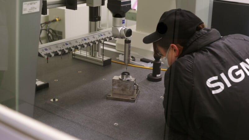A person wearing a jacket labeled "SIGN" is closely inspecting a metal component on a precision measuring machine inside a laboratory or industrial setting.