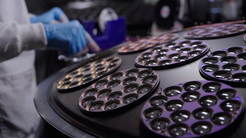 A person wearing blue gloves works near trays filled with multiple glass lenses, possibly in a laboratory or manufacturing environment. The trays are metallic and round, each holding several lenses.