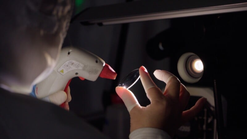 A person wearing gloves inspects a round glass lens under bright light, holding it carefully with one hand while using a handheld device in the other. A microscope or similar equipment is visible nearby.