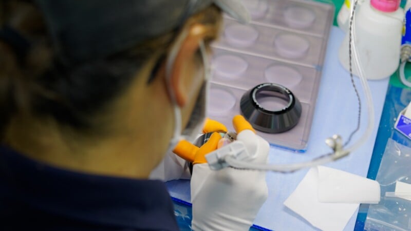 A person wearing gloves and a mask uses laboratory equipment at a workstation, handling a small object near a petri dish tray and various lab tools.