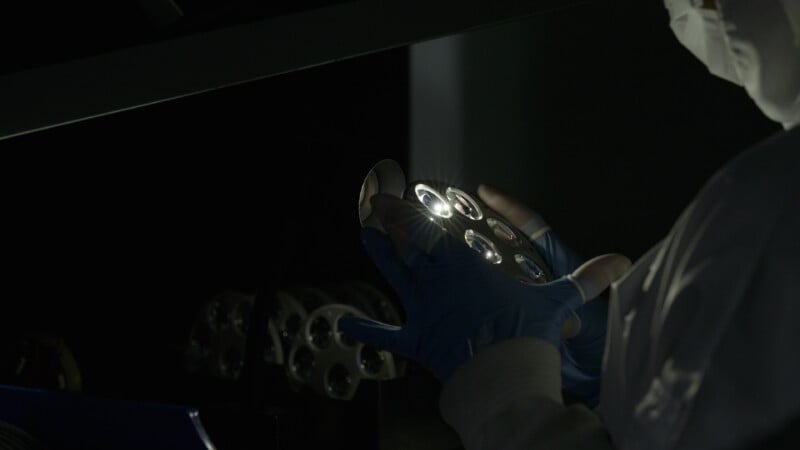 A person in protective clothing and blue gloves inspects a circular metallic object with multiple lenses or openings under focused light in a dimly lit environment.