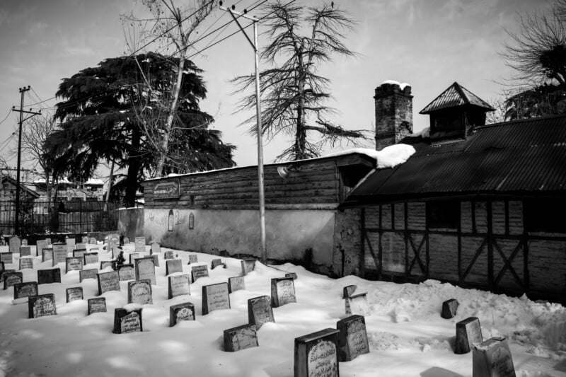 A snowy graveyard with numerous headstones stands beside a rustic wooden building under a cloudy sky. Tall trees and power lines are visible in the background. The scene is in black and white.
