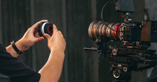 A person is adjusting a camera lens while positioned next to a professional video camera mounted on a tripod. The setup includes a large lens and an external monitor.
