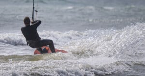 A person in a wetsuit rides a surfboard while kite surfing on choppy ocean waves, with water splashing around them.