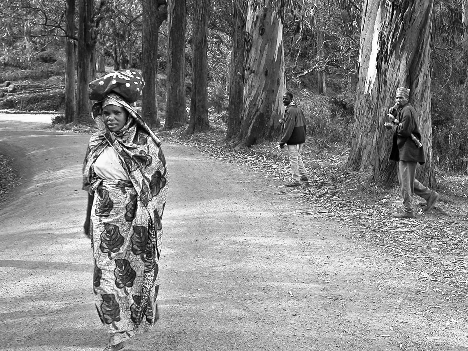 A woman in patterned clothing carries a bundle on her head while walking along a forested road. Two men stand near tall trees in the background, one looking back and the other holding a long knife over his shoulder. The image is in black and white.