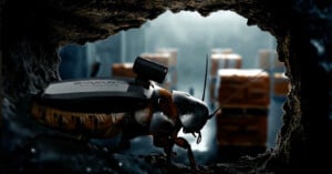 A close-up of a cockroach equipped with a small electronic device on its back, crawling through a hole, with blurred boxes visible in the background.