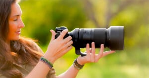 A woman with long brown hair holds a professional camera with a large telephoto lens outdoors, surrounded by green, blurred nature in the background.