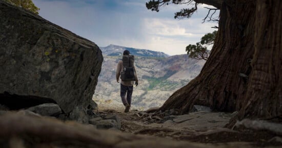 A hiker with a large backpack walks toward a scenic mountain vista, framed by a tree and a large rock, under a partly cloudy sky.