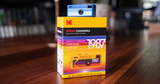 A blue Kodak Charmera keychain digital camera is placed on top of its colorful yellow and red box, which features bold "1987" text and graphics inspired by retro Kodak film packaging.