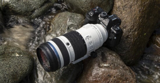 A black camera with a large white telephoto lens rests on wet rocks near flowing water, with visible water droplets on the camera and lens.
