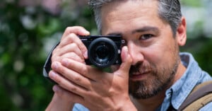 A man with gray hair and a beard is holding a camera up to his face, preparing to take a photo. He is outdoors, wearing a blue shirt, and the background is blurred greenery.