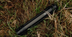 A black, collapsible camera tripod with carbon fiber legs lies folded on dry, patchy grass.