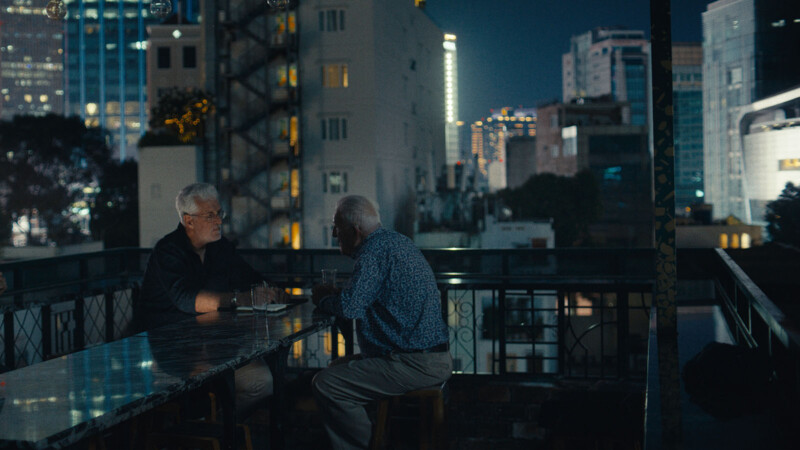 Two older men sit at a rooftop bar at night, engaged in conversation. City buildings and lights form the backdrop, creating an urban nighttime atmosphere. Drinks and glasses are on the marble table in front of them.