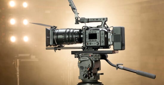 A professional video camera with a large lens and matte box mounted on a tripod, set up in a studio with bright lights in the background.