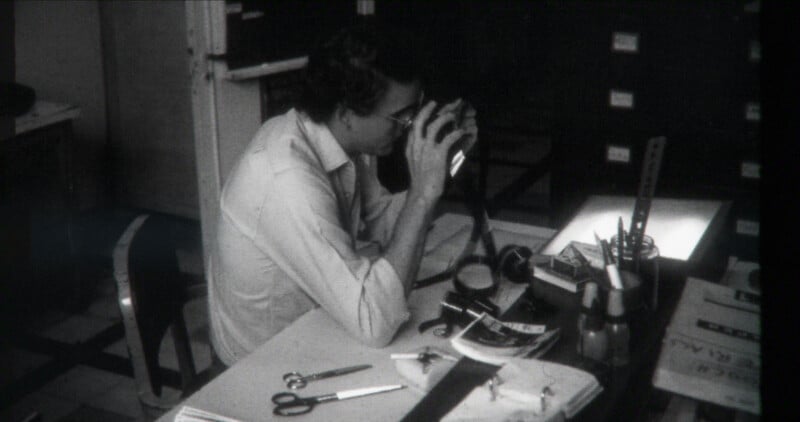 A person sits at a desk examining photographic film or slides with a magnifying device. The desk is cluttered with tools like scissors, rulers, and papers. The scene appears to be in an office or archival workspace.