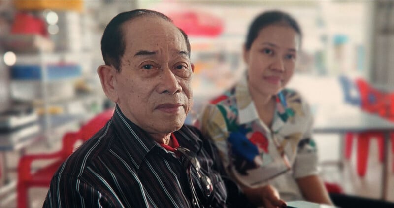 An older man in a dark striped shirt sits indoors next to a woman in a floral blouse. Both look toward the camera, with out-of-focus red and blue chairs in the background.