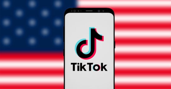 A smartphone displays the TikTok logo on its screen, positioned in front of a blurred American flag in the background.