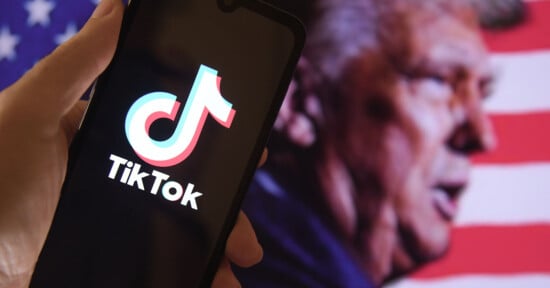A hand holds a smartphone displaying the TikTok logo, with a blurred image of a man’s side profile in front of an American flag in the background.