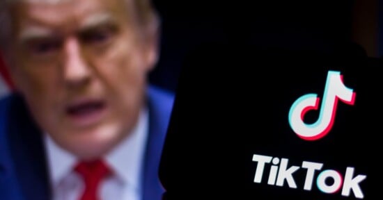 A close-up of a phone displaying the TikTok logo in white on a dark background. In the blurred background, there is an indistinct image of a person wearing a suit and red tie.