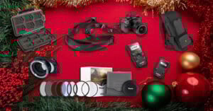 A festive holiday background with pine branches, baubles, and berries displaying camera accessories: memory card case, camera, lens adapter, filters, camera strap, remote controls, and a backpack.