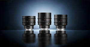 Three black camera lenses with "TTARTISAN" branding are displayed upright on a reflective surface against a blue-to-black gradient background, with dramatic lighting creating a professional look.