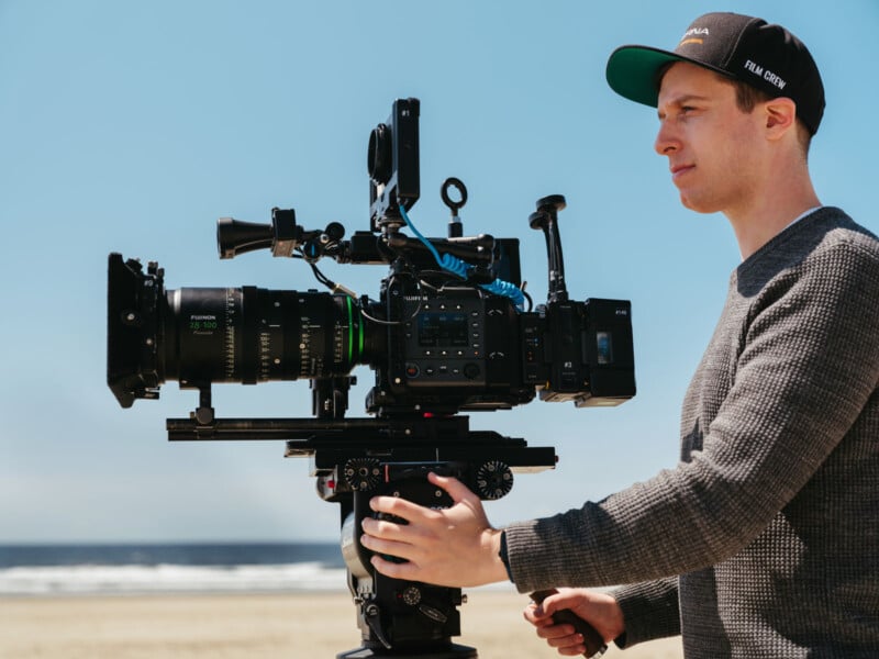 A person wearing a cap and sweater operates a professional video camera mounted on a tripod on a sandy beach, with the ocean and blue sky in the background.