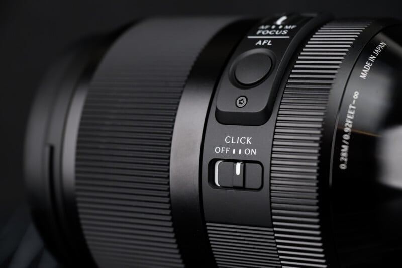 Close-up of a black camera lens showing buttons labeled "AFL," "CLICK OFF/ON," and focus settings, with textured grip details and "MADE IN JAPAN" visible on the lens barrel.